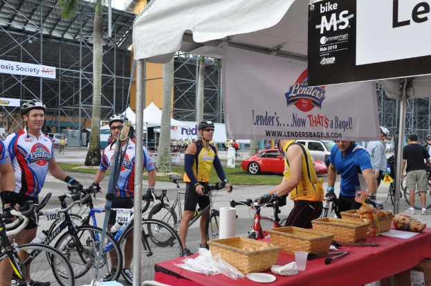 Eine Straßenszene mit Menschen, die in der Nähe von Fahrrädern stehen, einem Geschäft mit Gegenständen auf einem Tisch, Bäumen, Zelten, an eine Hütte angebrachten Schildern, Fahrzeugen und Gras.