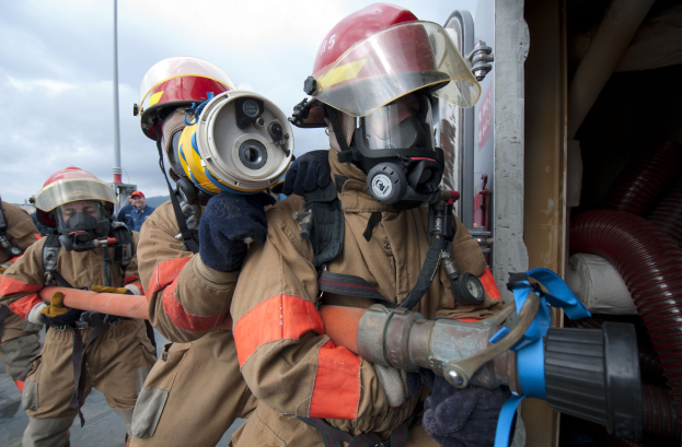 Feuerwehrleute in Schutzausrüstung mit einem Schlauch haltenden, neben einem Mast und Rohren vor einem bewölkten Himmel.