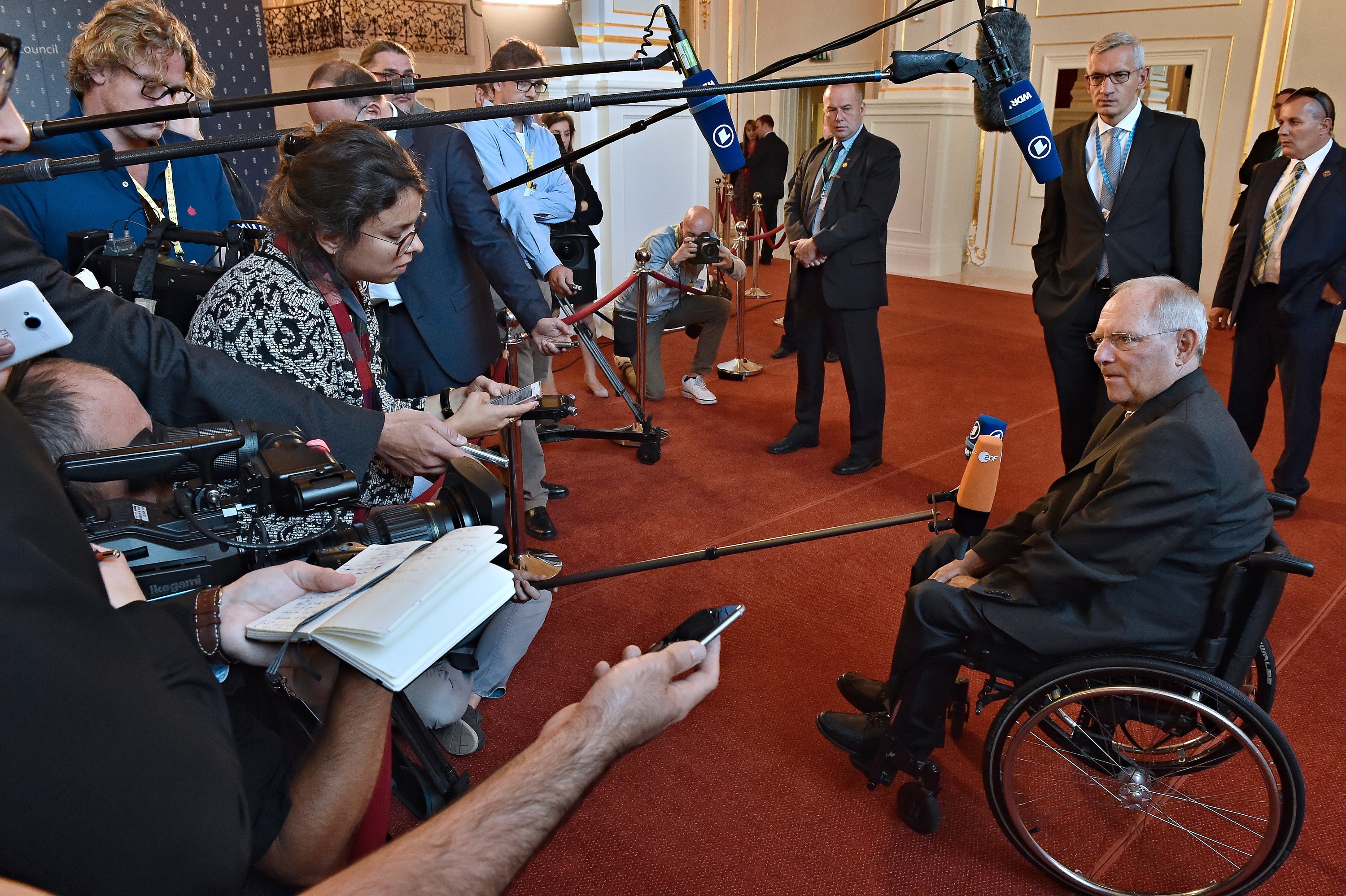 Ein Mann im Rollstuhl spricht mit Reportern, die Kameras, Mikrofone und Telefone halten, vor einem Banner, Scheinwerfern und einer Wand.