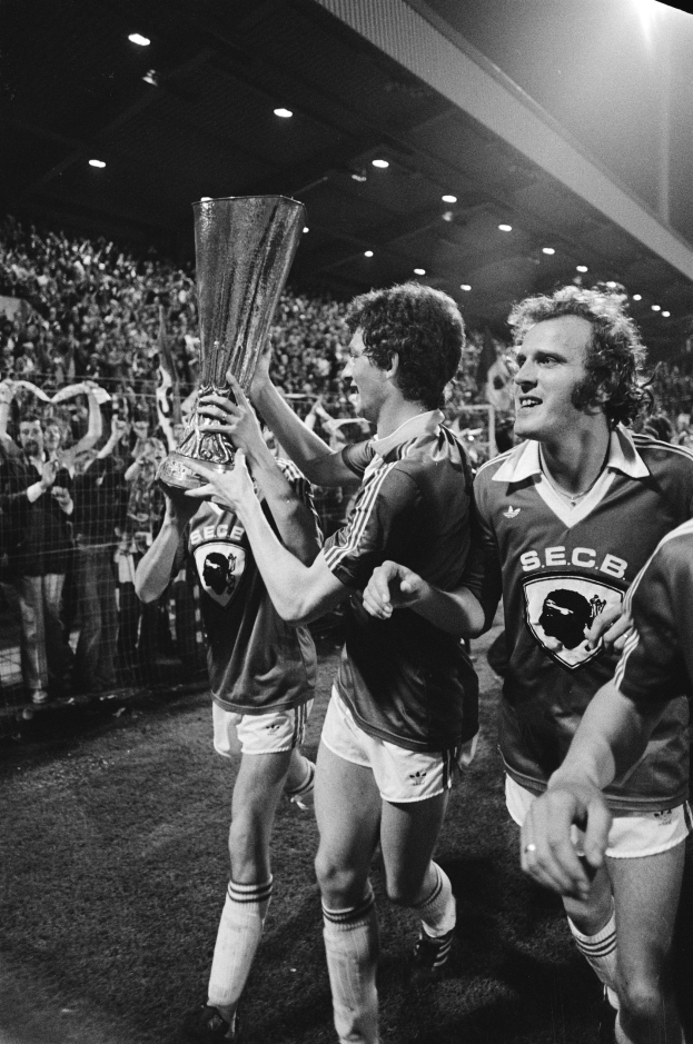 Schwarzes Foto von Fußballspielern, die mit einem Pokal auf einem Feld feiern, umgeben von einer Menge hinter einer Absperrung unter Stadionbeleuchtung.