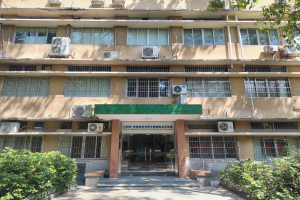 Ein großes Krankenhausgebäude mit zahlreichen Fenstern und Klimaanlagen, umgeben von Grünflächen, mit einer Straße, die darauf zuführt.