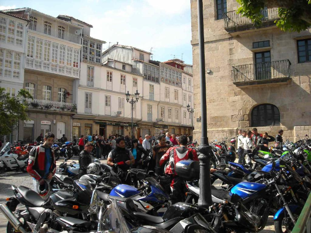 Ein cremefarbenes Gebäude mit zahlreichen Fenstern, das an ein Backsteingebäude mit einem Metallbalkon angrenzt, mit mehreren geparkten Fahrrädern und Menschen, die davor sprechen.