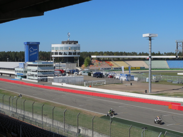 Eine Gruppe von Menschen auf Motorrädern auf einer Rennstrecke, umgeben von einem Zaun, Fahrzeugen, einem Gebäude, Schildern, Stadion und Bäumen unter einem bewölkten Himmel.