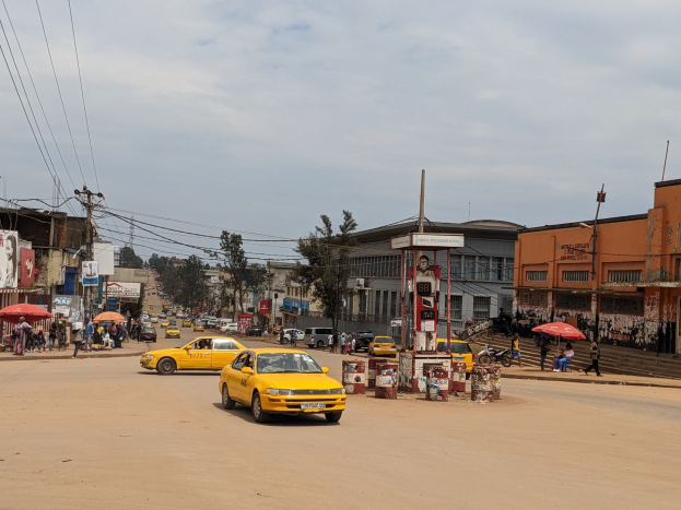 Ein gelbes Taxi fährt auf einer belebten Stadtstraße mit Gebäuden an beiden Seiten, während Fußgänger mit Regenschirmen auf dem Gehweg darunter gehen und der Himmel bewölkt ist.