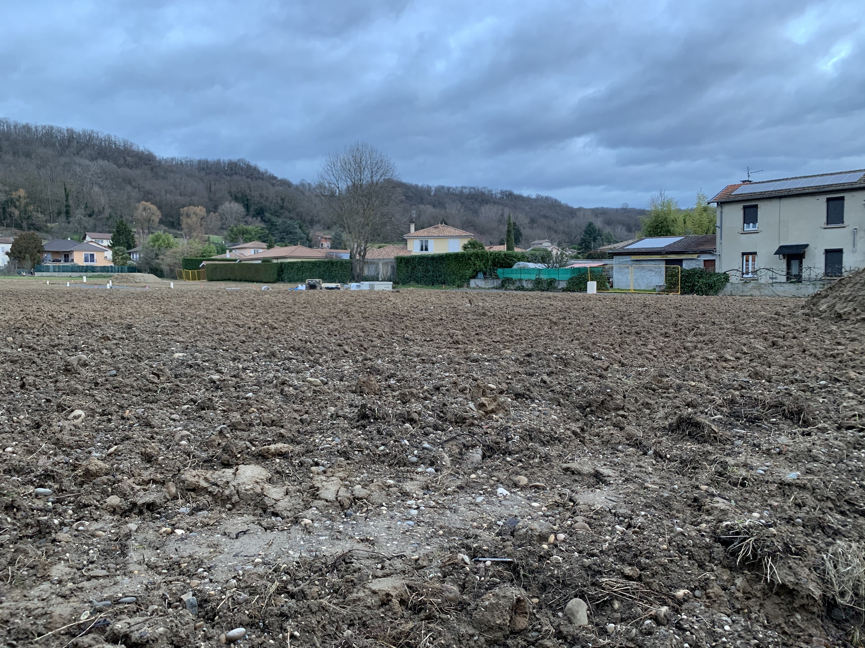 Ein großes freies Feld mit Erde, umgeben von Gebäuden, Bäumen, Pflanzen und einem Zaun, mit Hügeln und einer bewölkten Kulisse im Hintergrund; das Land ist zum Verkauf angeboten.
