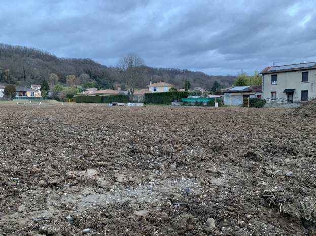 Ein großes freies Feld mit Erde, umgeben von Gebäuden, Bäumen, Pflanzen und einem Zaun, mit Hügeln und einer bewölkten Kulisse im Hintergrund; das Land ist zum Verkauf angeboten.