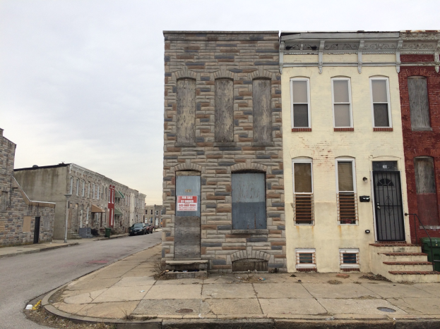 Ein altes, baufälliges Gebäude an einer Straßenkreuzung mit zerbrochenen Fenstern, bröckelnden Wänden, vorbeifahrenden Fahrzeugen, Mülltonnen auf dem Gehweg und einem klaren blauen Himmel im Hintergrund.