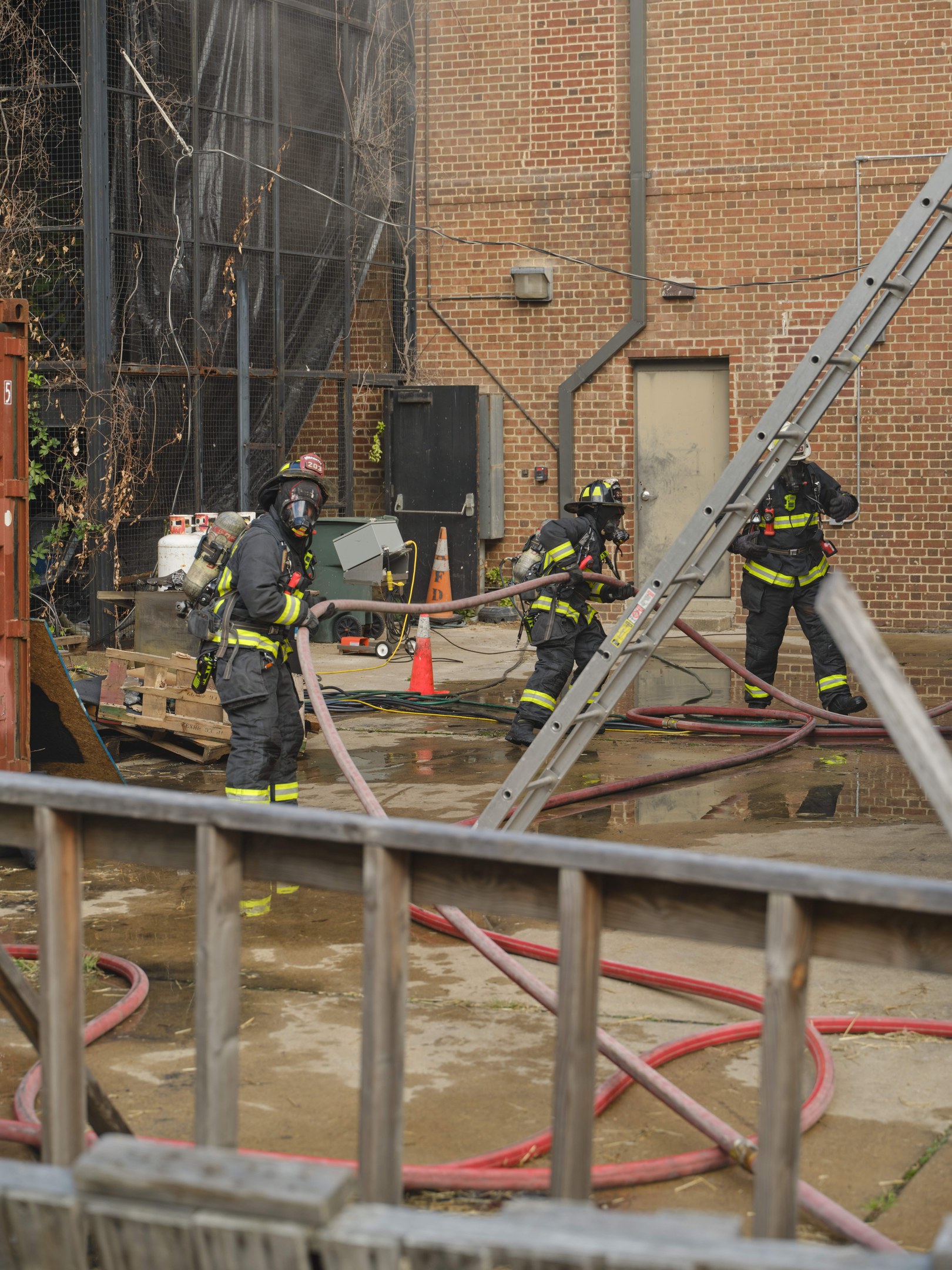 Feuerwehrleute in Helmen arbeiten daran, ein Gebäude Feuer zu löschen, umgeben von Equipment und einem Metallzaun.