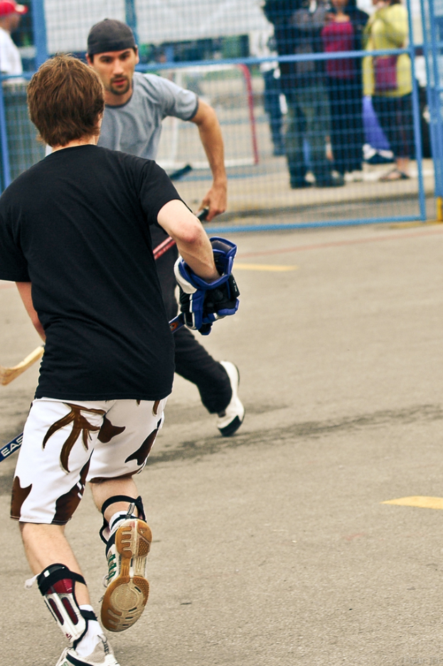 Zwei Personen spielen Hockey auf einer Straße mit einem Zaun und Zuschauern im Hintergrund.
