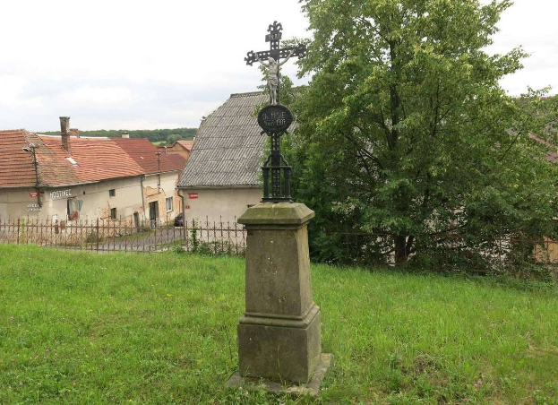 Ein hölzernes Kreuzdenkmal auf einer Wiese, umgeben von einem Metallzaun, mit Bäumen, Häusern und einem bewölkten Himmel im Hintergrund.
