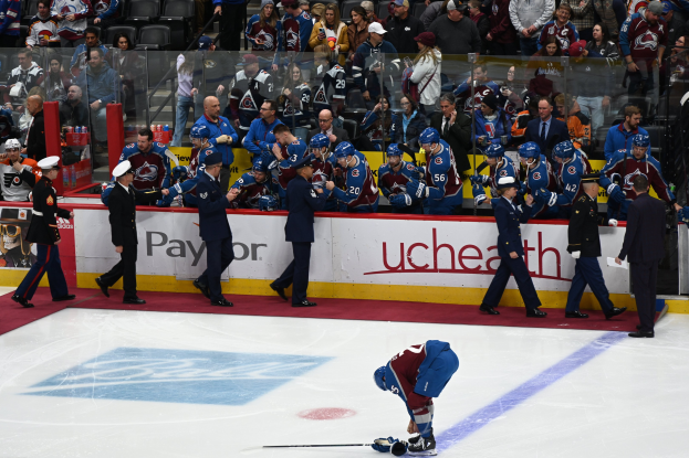 Ein Hockey-Spieler auf dem Eis umgeben von Teamkollegen und Gegnern, mit Zuschauern und Arena-Schildern im Hintergrund.