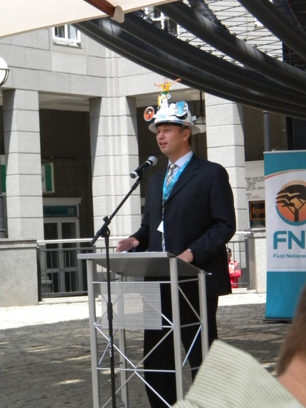 Ein Mann im Anzug und Hut spricht in ein Mikrofon auf einem Podium, mit einer Person rechts unten, einem Banner mit Text rechts und einem Gebäude mit Geländer im Hintergrund.