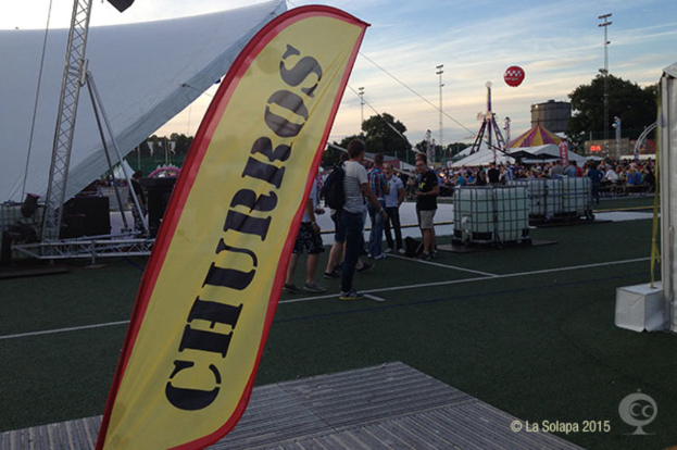 Faschingszelt mit Zelten, zentralem Mast, Menschen, Bäumen, Strommasten, Straßenbahnen auf der Straße und einem Eisenständer auf der linken Seite unter einem klaren Himmel.