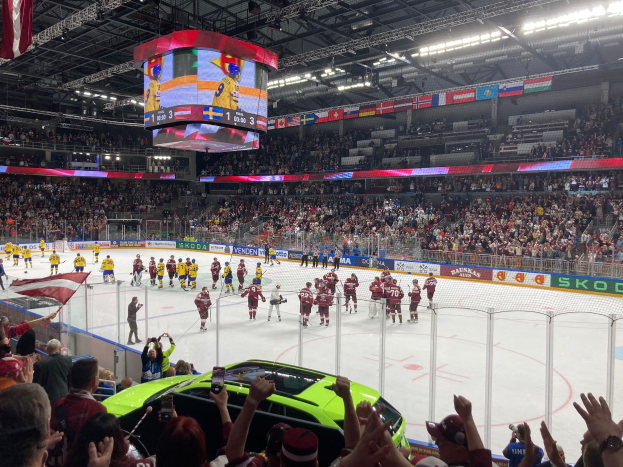 Hockey game in a large arena with spectators, rink fencing, banners, ceiling lights, a screen, and flags.