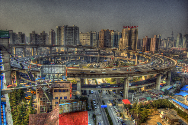 Eine Stadtansicht mit einer zentralen Ringstraße mit Fahrzeugen, einer unteren Straße mit Autos und Fahrrädern, zahlreichen Hochhäusern im Hintergrund, einem Himmel oben, Bäumen auf der linken unteren Seite und kleinen Häusern verteilt in der Mitte der Stadt.