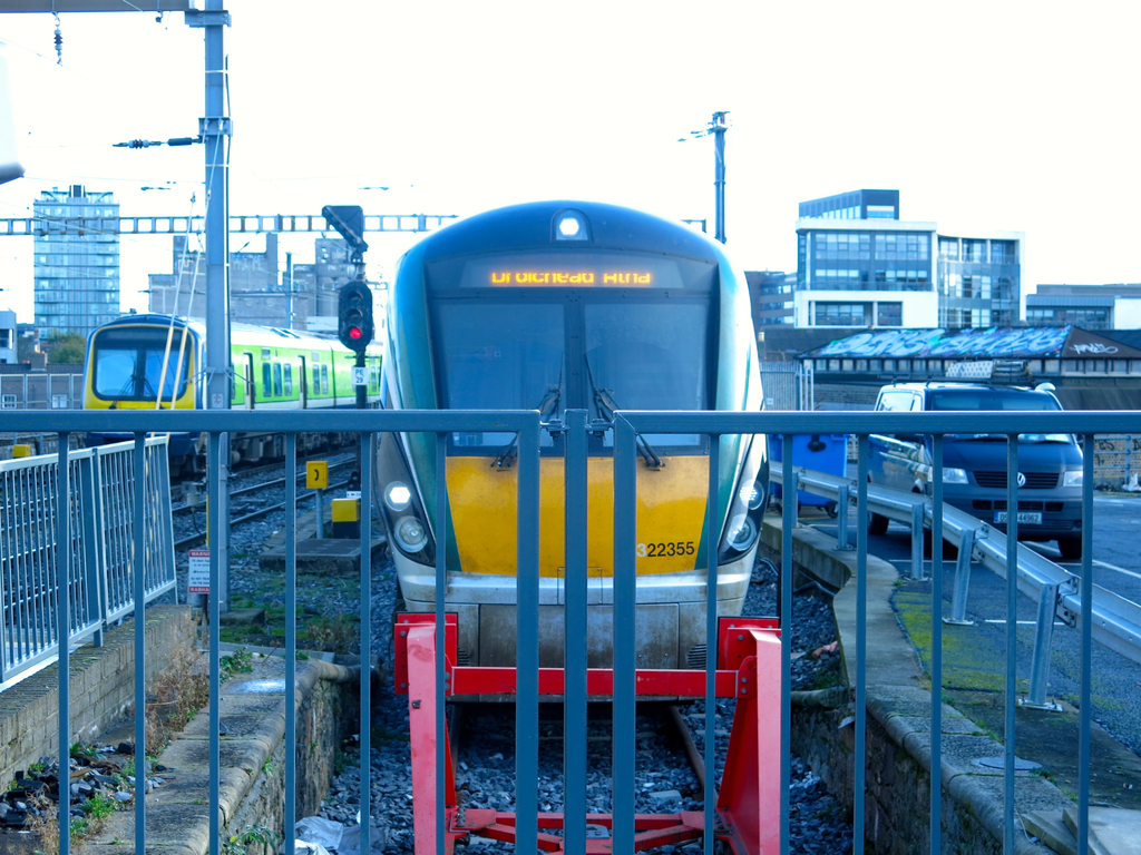 Zwei Züge befinden sich auf dem Gleis mit einem Zaun im Vordergrund, einigen Pfählen auf dem Land, einem mit einer Signalleuchte, einem Auto auf der Straße rechts, Gebäuden im Hintergrund und dem Himmel oben.