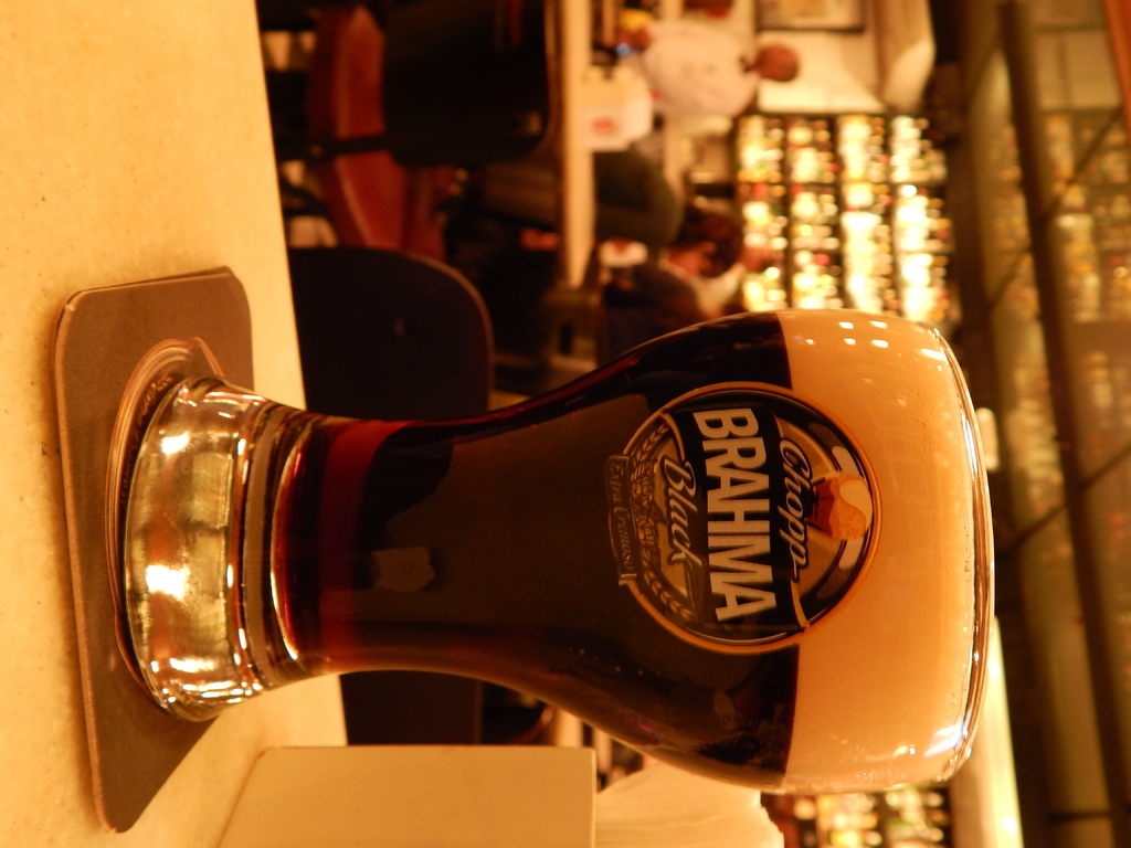 Ein Glas Wein auf einem Teller und eine Papiertaschentuchschachtel auf einem Weg, mit einer Gruppe von Menschen, Stühlen, Tischen, einem an der Wand befestigten Rahmen und Lichtern im Hintergrund.