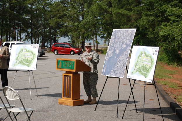 Eine Person steht an einem Podium mit einem Mikrofon, mit Plakaten, Stühlen, Fahrzeugen und Bäumen im Hintergrund.