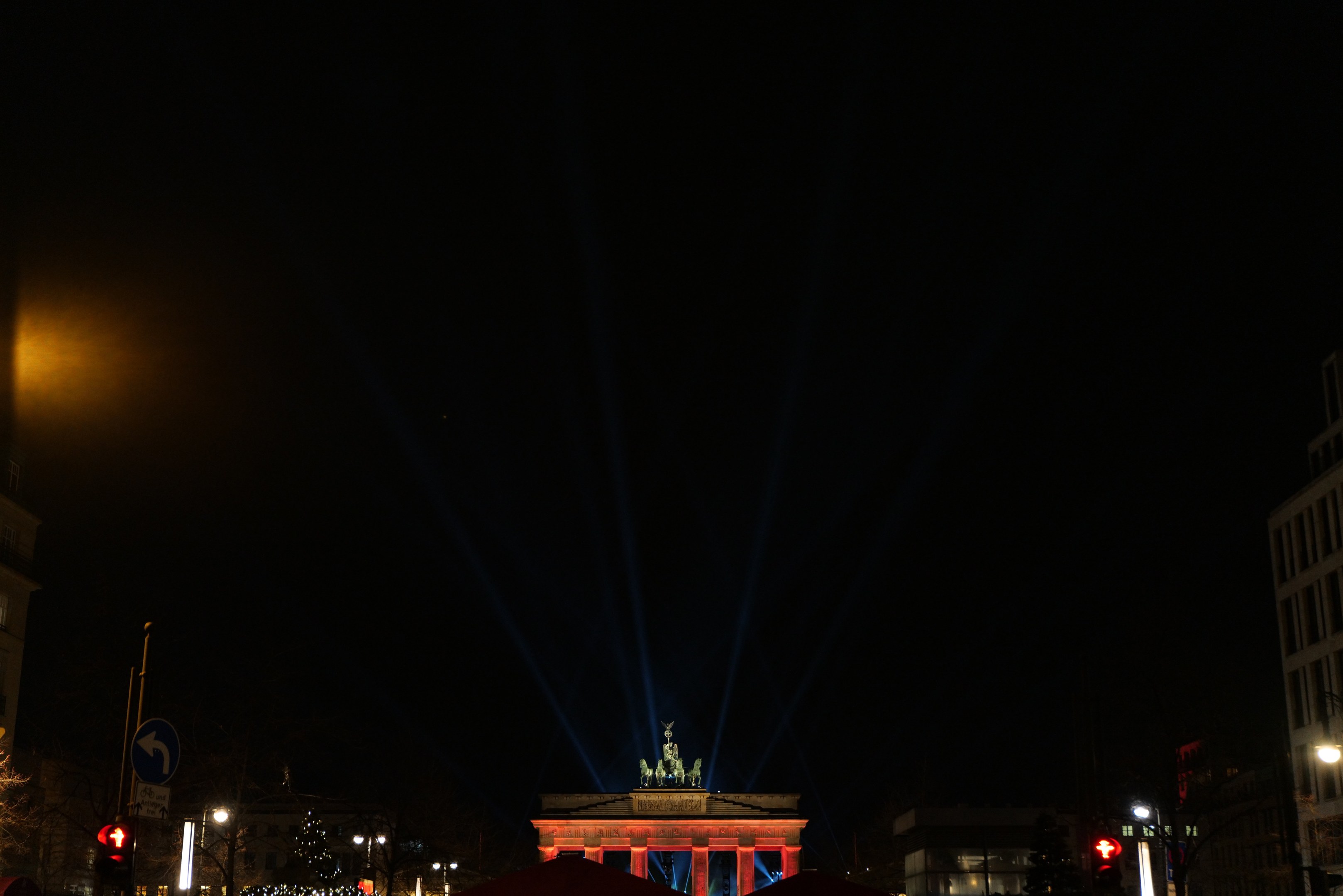 Brandenburger Tor bei Nacht, von Straßenlaternen beleuchtet, umgeben von Gebäuden, Bäumen, Pfählen, Verkehrszeichen, Schildern und Zelten unter einem dunklen Himmel.