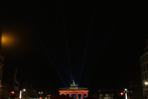 Brandenburger Tor bei Nacht, von Straßenlaternen beleuchtet, umgeben von Gebäuden, Bäumen, Pfählen, Verkehrszeichen, Schildern und Zelten unter einem dunklen Himmel.