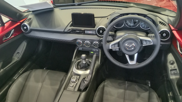 Innenansicht eines roten Mazda MX-5 Miata RF mit schwarzen Ledersitzen, Lenkrad, Schaltstange, Knöpfen, Bildschirm, Spiegel und anderen Innenraumkomponenten.