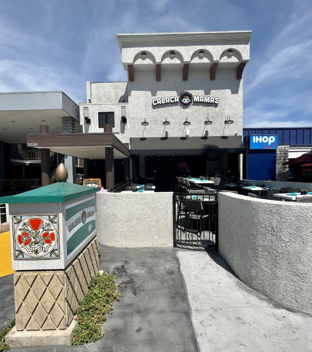 Außenansicht des Calaca Mama's-Restaurants in San Diego, Kalifornien, mit Tischen und Stühlen vor einer Mauer mit einem Tor, Pflanzen mit Blumen, einer Tafel an der Wand und einem bewölkten Himmel im Hintergrund.
