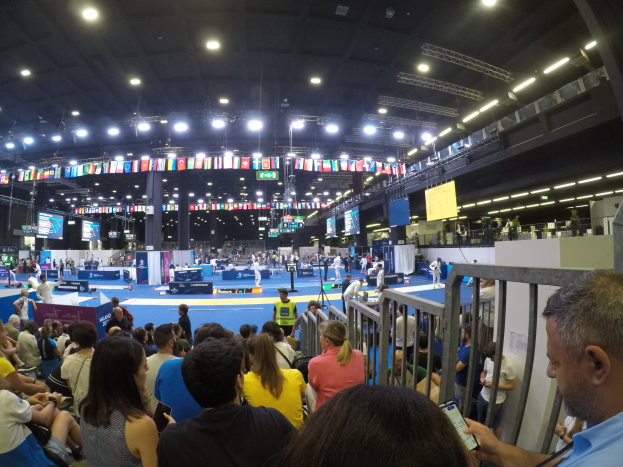 Eine große Menschenmenge sitzt und steht in einer Indoor-Sportarena, mit Metallzaun auf der rechten Seite, Bildschirmen, Flaggen und Deckenbeleuchtung im Hintergrund.
