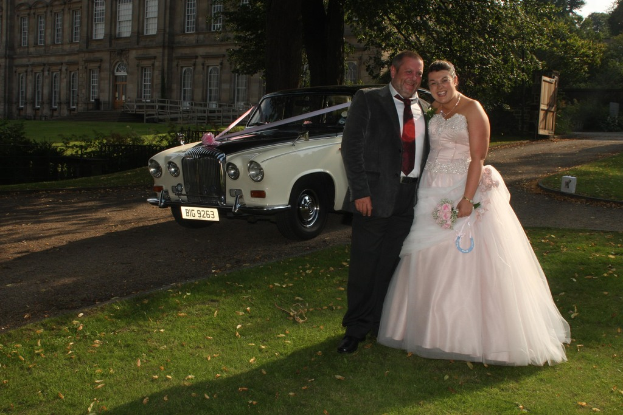 Eine Braut und ein Bräutigam stehen zusammen, einer hält Blumen und ein Objekt, mit Bäumen, einem Gebäude, Geländern, einem Tor und einem Fahrzeug im Hintergrund.