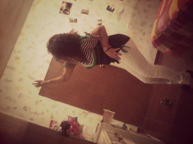 Eine Frau steht mit einer Kamera in der Hand, trägt ein schwarzes Oberteil und weiße Jeans, vor einer weißen Wand und einer braunen Tür und einem Bett im oberen rechten Bildrand.