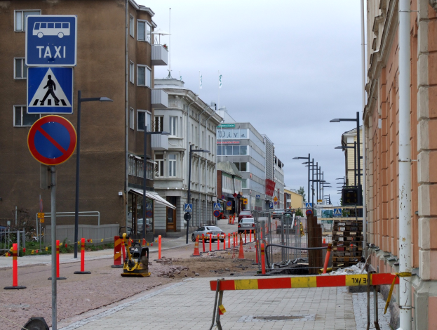 Stadtstraße mit Gebäuden, Laternenmästen, Lichtern, Schildern, Verkehrskegeln, Fahrzeugen, Absperrpollern, Bäumen, einer Baustelle mit Verkehrsschildern und einem bewölktem Himmel.