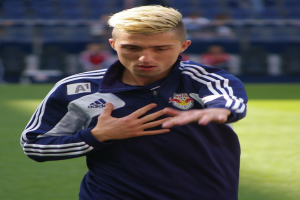 Ein junger Mann in einem blauen T-Shirt steht auf einem Fußballfeld und streckt die Arme aus, wobei er entschlossen wirkt und im Hintergrund unscharf ist.