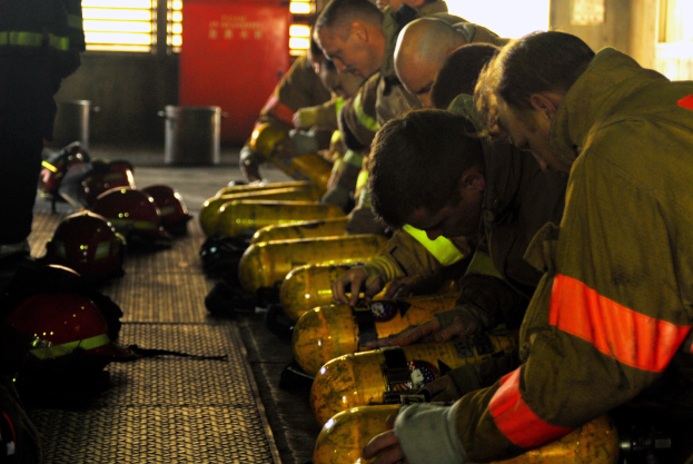 Feuerwehrleute in Schutzausrüstung arbeiten in der Nähe eines Feuerlöschfahrzeugs mit Zylindern und Helmen auf dem Boden, in einem Raum mit Fenstern, einer Tür und einem Müllcontainer.