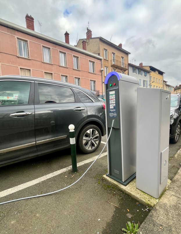 Ein Elektroauto ist an einer Ladesäule am Straßenrand eingesteckt, mit Gebäuden, Laternenmästen und einem bewölkten Himmel im Hintergrund.