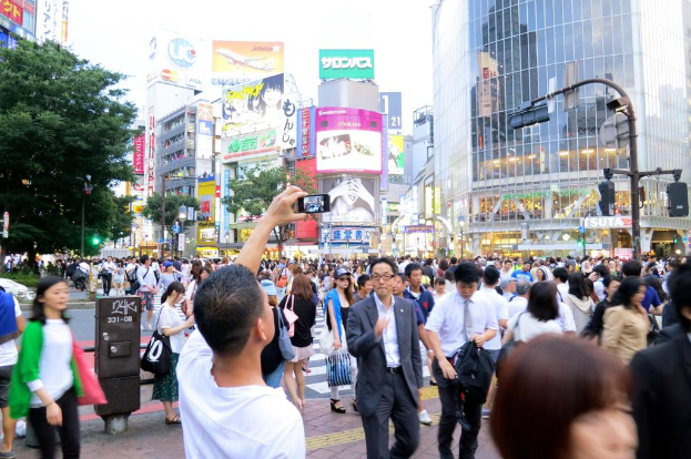 Eine Gruppe von Menschen, die auf einem Weg gehen, wobei ein Mann ein Mobiltelefon hält, umgeben von Bäumen, Pfählen, Verkehrszeichen, Gebäuden und Schildern mit Text.