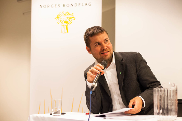 Eine Person in einem Anzug lächelt, hält ein Mikrofon und ein Dokument in der Hand und steht an einem Tisch mit einem Glas und einem Handy, mit einer weißen Fahne und einer Wand im Hintergrund.