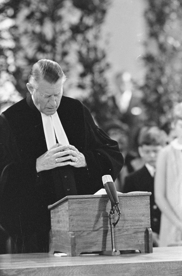 Ein Mann in einem schwarzen und weißen Gewand steht an einem Podium mit einem Mikrofon und spricht zu einer kleinen Gruppe von Menschen mit Bäumen im Hintergrund.