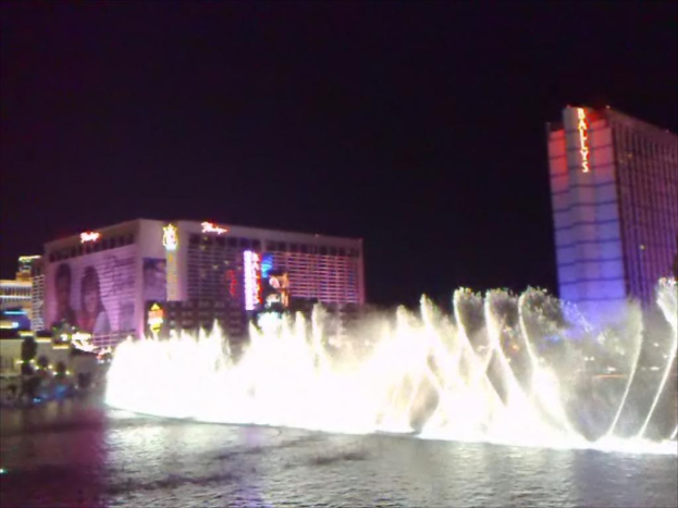 Ein Springbrunnen mit Wasser an seiner Basis, umgeben von Gebäuden, Plakaten, Bildschirmen, Lichtern, Pflanzen und Himmel.