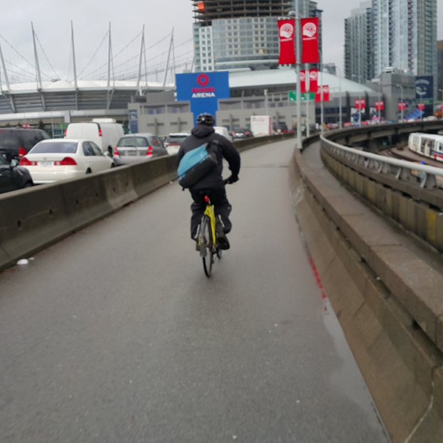 Eine Person mit Helm und Rucksack sitzt auf einem Fahrrad auf einer Straße mit parkenden Autos und Gebäuden im Hintergrund, mit Bannern auf nahegelegenen Pfosten.