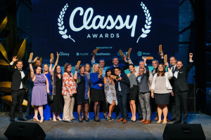 Eine Gruppe von Menschen auf einer Bühne, die lächeln und Auszeichnungen halten, mit Lautsprechern unten und einem Bildschirm im Hintergrund, der "Classy Awards 2019" anzeigt.