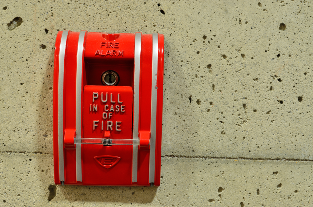 Ein roter, zylindrischer, metallener Brandmeldekasten mit einer glänzenden Oberfläche, einem Griff oben, einem Schlüsselloch in der Mitte und der Aufschrift "Brandmeldeanlage" in fetter schwarzer Schrift, an der Seite eines Gebäudes montiert.