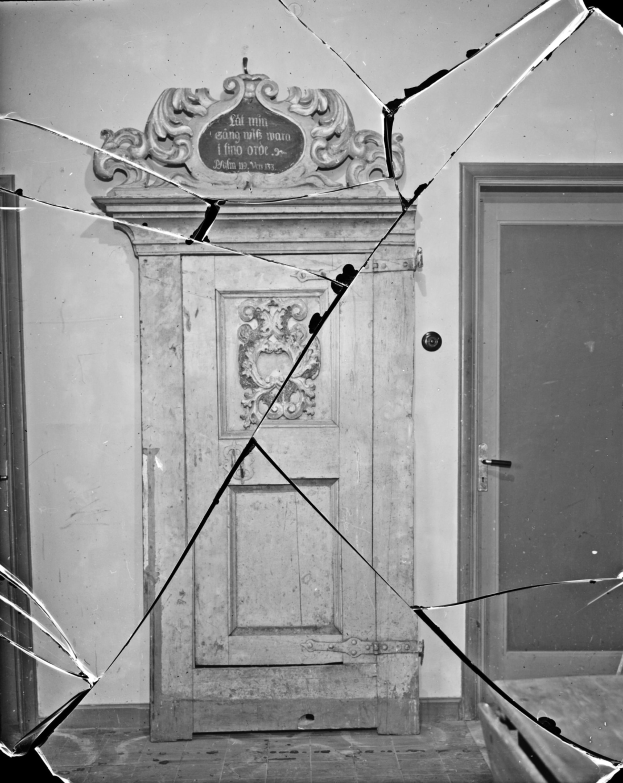 Schwarz-weiß-Foto einer zerbrochenen Tür in der Mitte eines Raumes mit Glasscherben drumherum und einer Wand im Hintergrund.