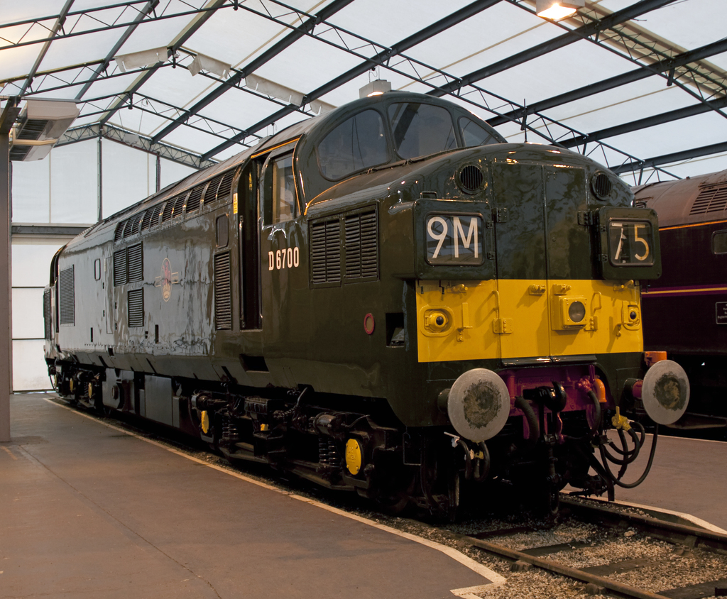 Ein Zug fährt auf den Schienen neben einem Bahnsteig mit einem Mast auf dem Dach, der Metallstangen mit Lampen trägt.