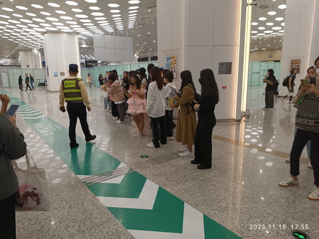 Eine Gruppe von Menschen steht in einer Reihe an einem Flughafen-Check-in, einige halten Taschen und Mobiltelefone, mit Säulen im Hintergrund, Lampen an der Decke und einem Wasserzeichen am unteren Bildrand.