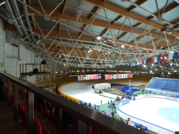 Großes Indoor-Eisstadion mit zahlreichen Menschen auf der Eisfläche, umgeben von Sitzplätzen, Geländern, Brettern, Beleuchtungskörpern und konstruktiven Elementen, mit einer Wand im Hintergrund.