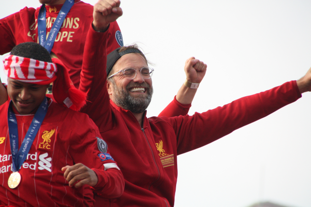 Liverpool-Trainer Jürgen Klopp feiert in rotem Jackett mit seinen Spielern nach dem Gewinn der UEFA Champions League, umgeben von zwei medaillentragenden Personen und einem Himmel im Hintergrund.