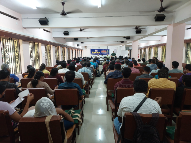 Eine große Gruppe von Menschen sitzt in Stühlen in einem Raum mit Fenstern an den Seiten, Säulen hinten, einem Banner in der Mitte und Ventilatoren und Lampen an der Decke und nimmt an einem Workshop zum Thema Bedeutung der Bildung in Indien teil.