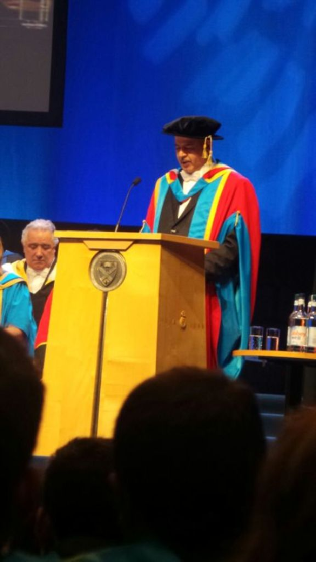 Ein Mann in einer Graduationsrobe und -kappe steht an einem Podium mit einem Mikrofon, mit Menschen, die darunter und links sitzen, und einem Tisch mit Gl├Ąsern und Flaschen rechts, vor einem blauen und schwarzen Hintergrund.