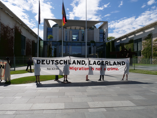 Eine Gruppe von Menschen hält ein Transparent mit der Aufschrift "Deutschland, Lagerland Migration ist kein Verbrechen" vor einem Gebäude mit Fenstern, umgeben von Gras, einem Metallzaun, Pflanzen, Bäumen und Fahnenmästen, unter einem bewölkten Himmel.