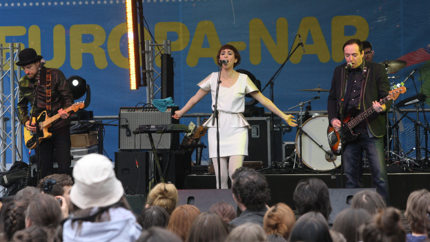 Eine Konzertaufführung mit vier Personen auf der Bühne: eine Frau in einem weißen Kleid, die in der Mitte singt, zwei Männer, die Gitarren auf beiden Seiten spielen, und eine Menge vor einer Wand mit Lautsprechern im Hintergrund.