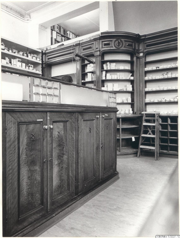 Schwarzes und wei├čes Foto eines vintage Apothekeninneren mit einer Theke, Sch├Ąben, einer Leiter, Regalen voller Beh├Ąlter und Text überall.